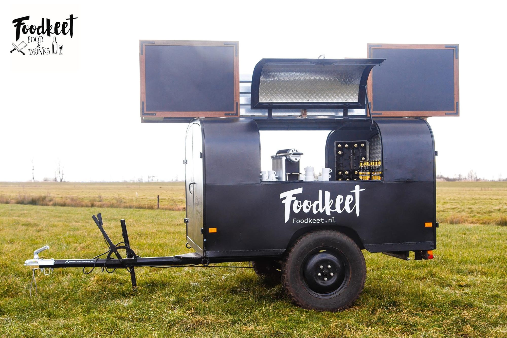 Zwarte Foodkeet-aanhanger in een weiland, met open luiken, een koffiemachine, kopjes en flesjes bier